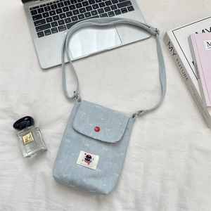 Detail van lichtblauwe denim crossbodytas met rode knoop en kattenpatch op witte textielstof