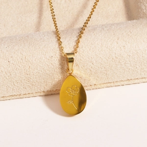 Collier élégant en acier doré avec pendentif goutte gravé de fleur sur un support beige