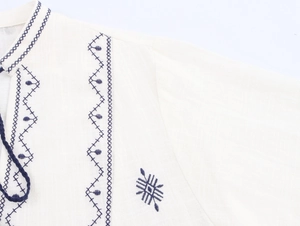 Detail of blue geometric embroidery and woven drawstrings on a white cotton linen blouse