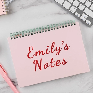 Detail of pink spiral notebook featuring custom red glitter lettering on marble desk