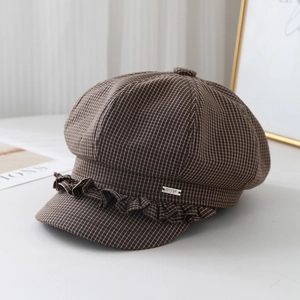 Detail of a brown plaid newsboy cap featuring ruffle trim and a silver metal logo plaque
