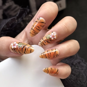 Detailed view of almond faux nails featuring orange tiger stripes and 3D floral accents
