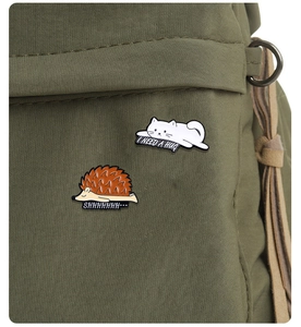 Close-up of cute enamel cat and hedgehog pins on green canvas with 'I NEED A HUG' text