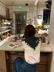 Detail of dark green knitted triangle scarf with fringe worn by a model in a cozy cafe