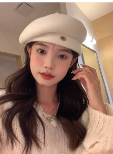 Detailed view of an ivory wool beret featuring an oval metallic letter M ornament
