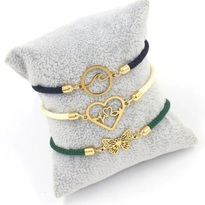 Detailed view of woven bracelets with gold wave, heart, and butterfly charms on a grey pillow