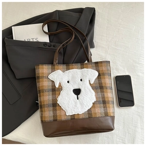 Brown plaid tote bag with plush dog patch shown in a stylish lifestyle setting on a bed