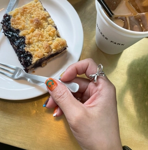 Close-up of a silver bow ring worn on a hand in a cafe setting with coffee and cake