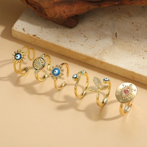 Flat lay of six gold rings featuring evil eye, sun and floral motifs on a stone slab