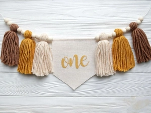 Detail of fabric first birthday banner with golden one text, colorful tassels and wood beads