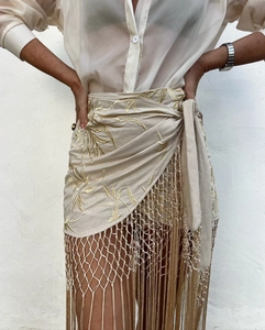 Détail de mini-jupe portefeuille beige avec broderie dorée et longues franges tressées