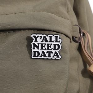 Close-up of enamel pin with Y'ALL NEED DATA text on olive green canvas fabric
