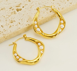 Close-up of golden geometric twist earrings with white pearls on a light stone surface