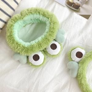 Close-up of green plush frog headband with large 3D eyes on a white soft surface
