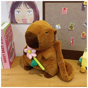 Brown plush capybara crossbody bag holding a pink flower on a wooden desk setup