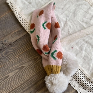 Detail showing pink knitted scarf with orange flower patterns and white faux fur pom poms