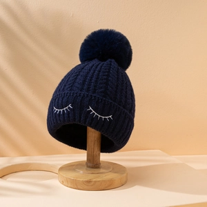 Dettaglio del berretto in maglia blu scuro con pompon e ricamo ciglia giocoso