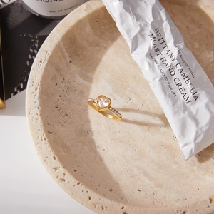 Dettaglio anello a cuore in acciaio oro 18K con zirconi su vassoio decorativo in travertino