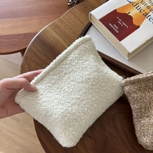 Beige tweed makeup bag with woven texture on a wooden table next to a book