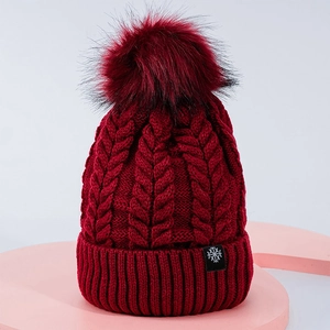 Close-up of wine red cable knit beanie with fluffy pom-pom and snowflake logo patch