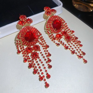 Detailed close-up of gold fringe earrings with red crystals on a textured beige tray
