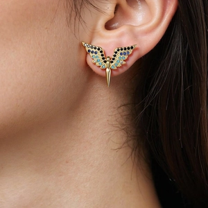 Close-up of golden wing-shaped earrings with blue zircon stones on a model's ear