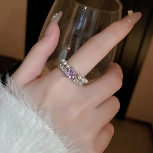 Gros plan d'une bague cœur avec zircon rose et perles portée à la main sur un verre