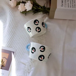 Top-down view of ceramic panda mugs with blue handles and hand-painted art on white fabric