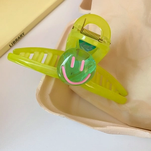 Détail de la pince à cheveux verte avec motif smiley rose sur plateau et livre jaune