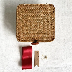Detail of woven straw gift box with red ribbon, thank you tag, and pearl pin on white linen