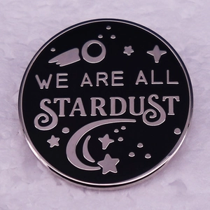 Black enamel pin with silver 'WE ARE ALL STARDUST' text and astronomical patterns