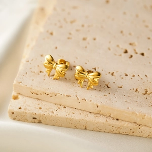 Close-up of golden ribbon bow earrings on beige travertine stone with soft white fabric