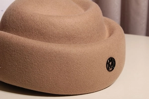 Detailed macro view of camel wool felt hat featuring a metallic M logo ornament