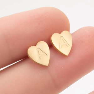 Détail d'une boucle d'oreille cœur dorée avec une lettre A gravée tenue entre les doigts