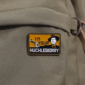 Detail of I'm Your Huckleberry enamel pin displayed on a green canvas tactical backpack