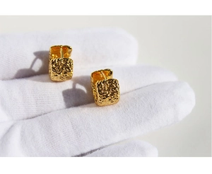 Detailed view of square gold earrings with unique lava texture held in white gloves