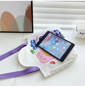 White canvas tote bag with purple straps and cartoon print shown next to a tablet for scale