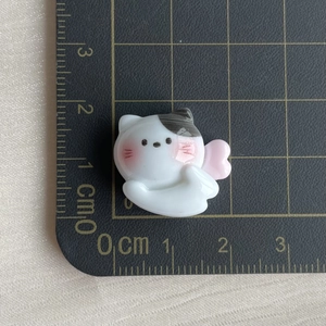 Detail shot of a white resin cat charm with pink wings on a metric scale background