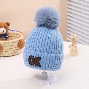 Detail of light blue knit kids hat with thick grey faux fur pompom and OK badge