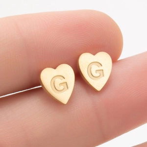 Gros plan d'une boucle d'oreille cœur en or avec gravure de la lettre G entre les doigts