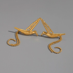 Détail de clous d'oreilles colibri dorés avec de fines chaînes perlées sur fond neutre