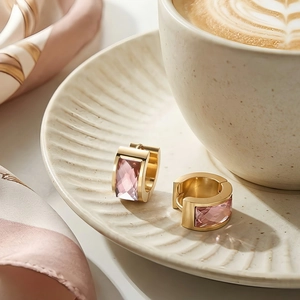 Detail of gold U-shaped earrings featuring rectangular pink crystal on white ceramic plate