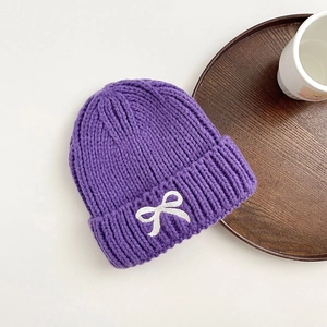 Close-up of purple knit beanie featuring a white embroidered bow and thick cuffed texture