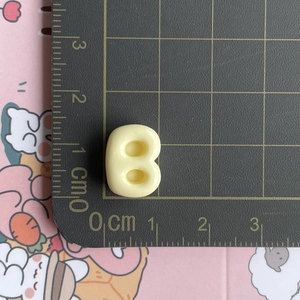 Detailed macro view showing a beige resin letter B accessory on a centimeter scale mat