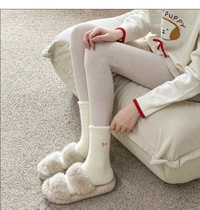 Detail of white plush socks with pink bow embroidery and beige slippers in home setting