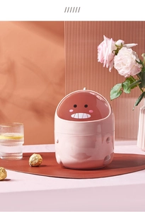 Pink dinosaur desk trash bin with flip lid shown in a cozy office setting