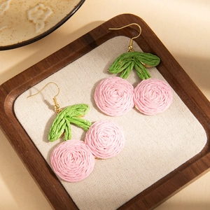 Macro detail of handmade woven cherry earrings with pink globes and green leaves in a tray