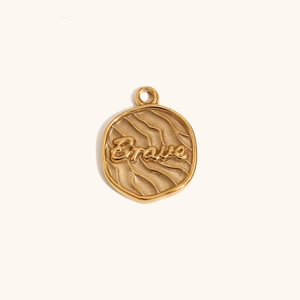 Macro detail of round 18K gold plated pendant with Brave engraving and wavy texture