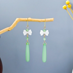 Detail of long drop earrings featuring a white bow and green teardrop pendant on wood