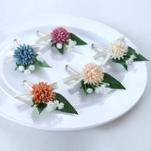 Five assorted wedding boutonnieres with artificial flowers and silk ribbons in a ceramic tray